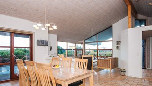 Un comedor amplio con una mesa y sillas de madera, ventanas grandes que ofrecen vistas a la vegetación y un techo con vigas de madera a la vista.