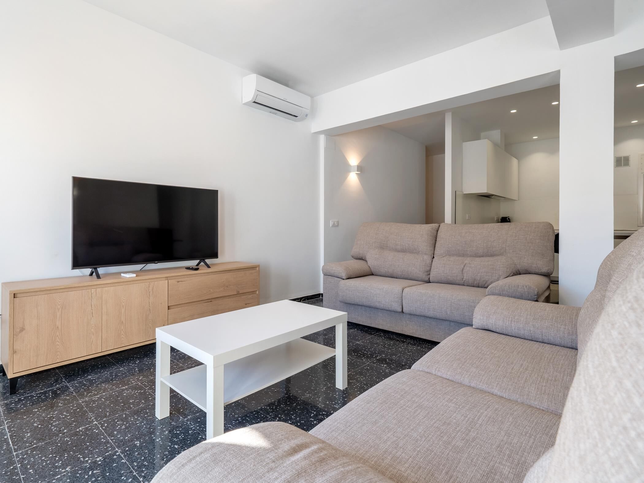 Una sala de estar moderna con un televisor de pantalla plana, un mueble de madera, una mesa de centro blanca y un sofá color beige.