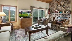 A cozy living room with a stone wall, wooden ceiling, and a television displaying a golf scene. - 100 Ridgepoint 4 Bedroom Condo (Avon)