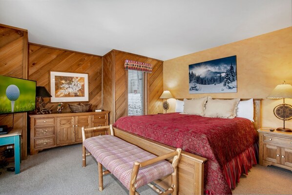 A bedroom with a wooden headboard, a large bed with a red blanket, a wooden bench, a dresser, a television, and two wall-mounted lamps.