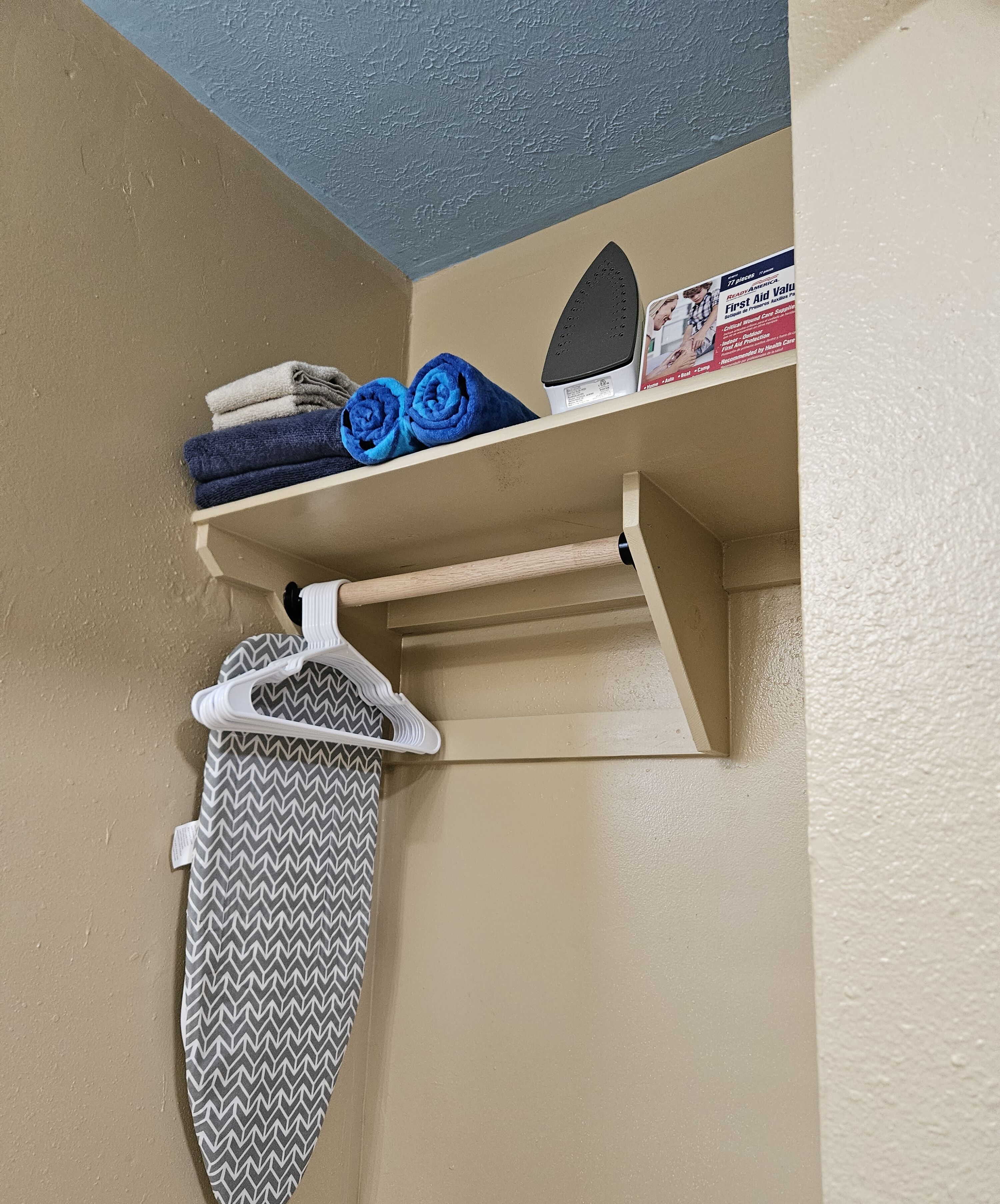 A corner of a room with a beige wall, a blue ceiling, and a shelf with folded towels and a first aid kit.