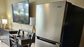 A compact kitchenette with a stainless steel refrigerator, a dining table with chairs, a TV mounted on the wall, and a small cabinet.