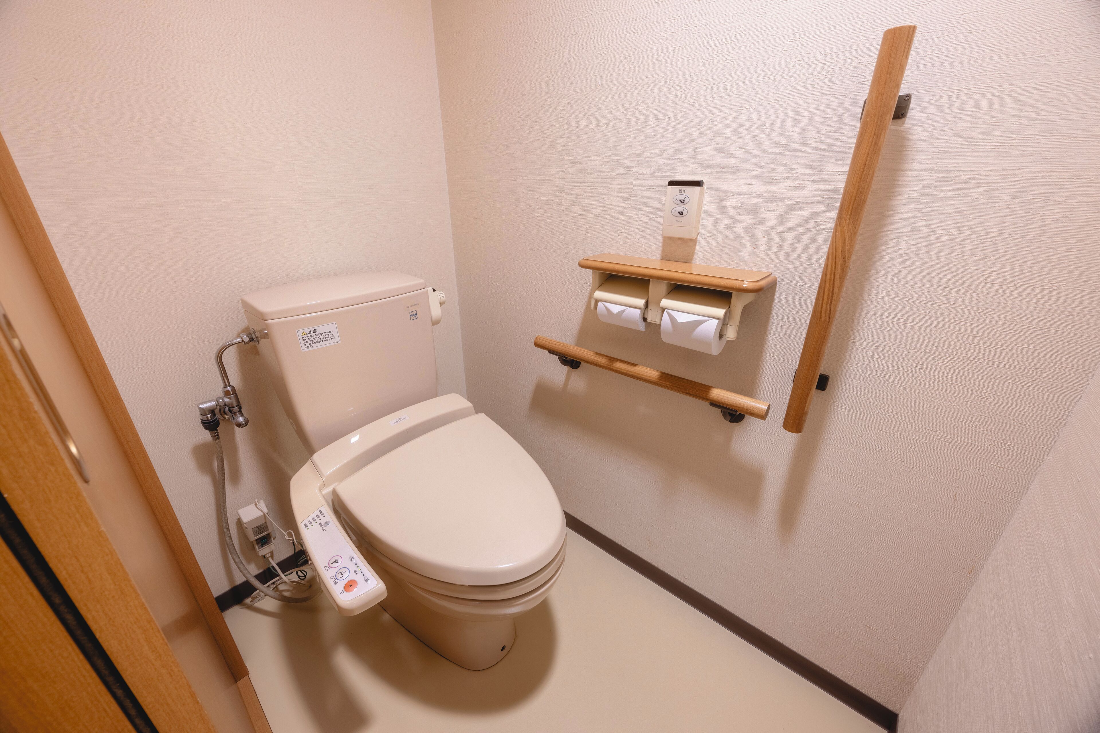 A standard toilet with a control panel, a wooden shelf with toilet paper, and a wooden handrail.