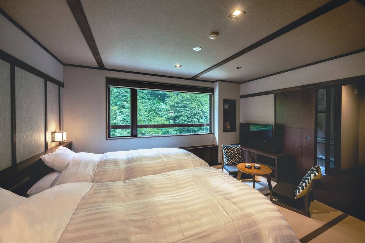 Une chambre d’hôtel avec deux lits, une télévision, une petite table et une fenêtre donnant sur un paysage verdoyant.