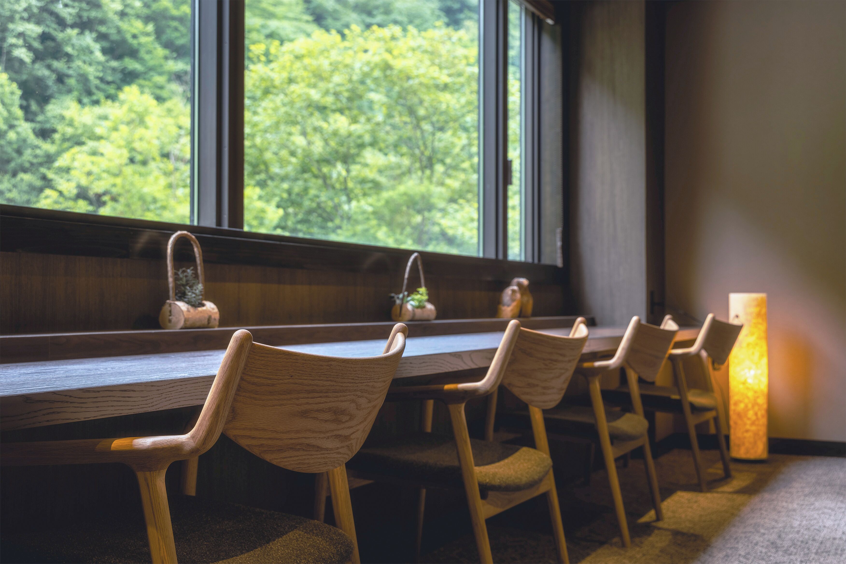 A dining area with a long wooden table and chairs, a window with a view of trees, and a tall, textured floor lamp.