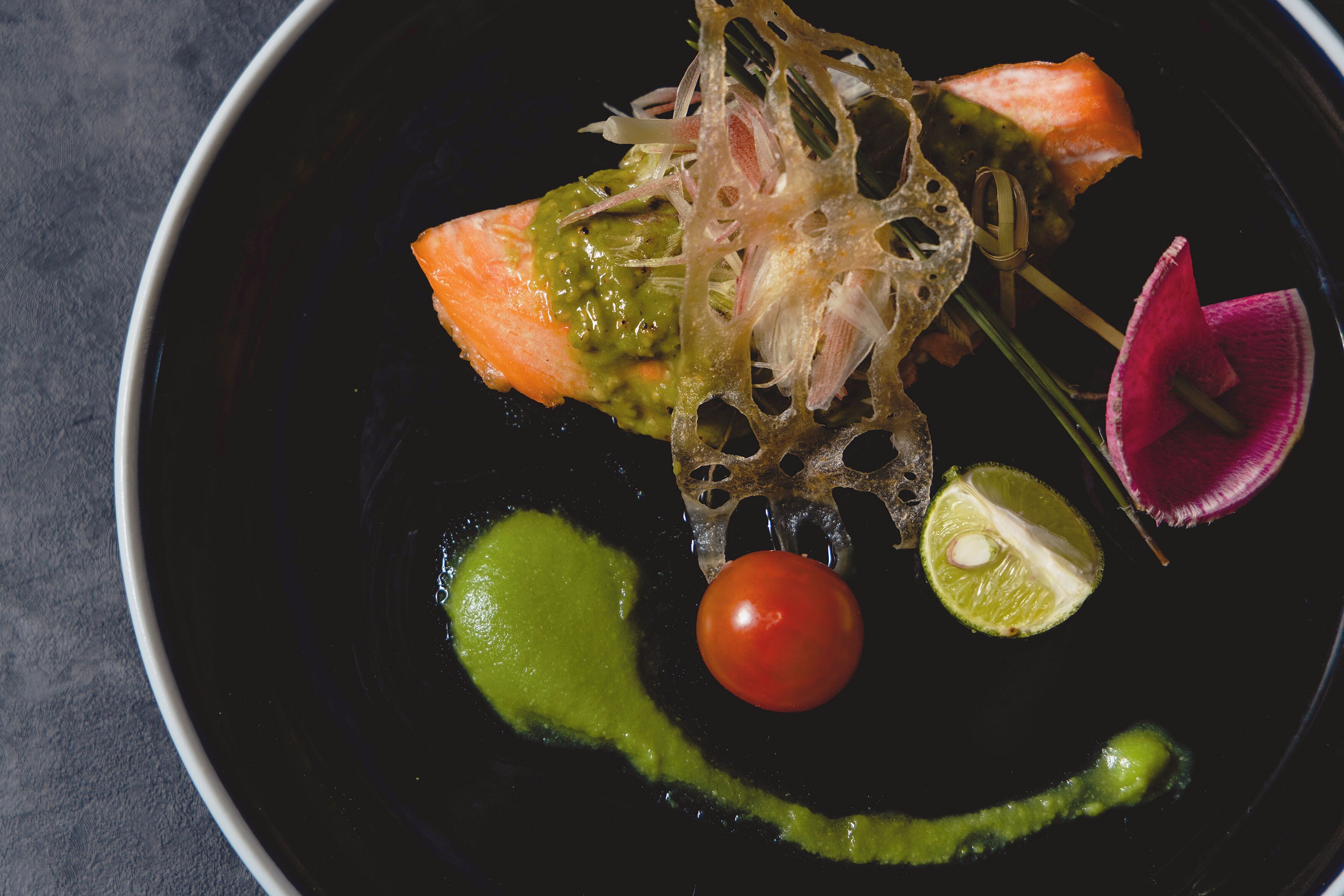 Une assiette noire avec un morceau de saumon, une sauce verte, une tomate cerise et une tranche de citron vert.
