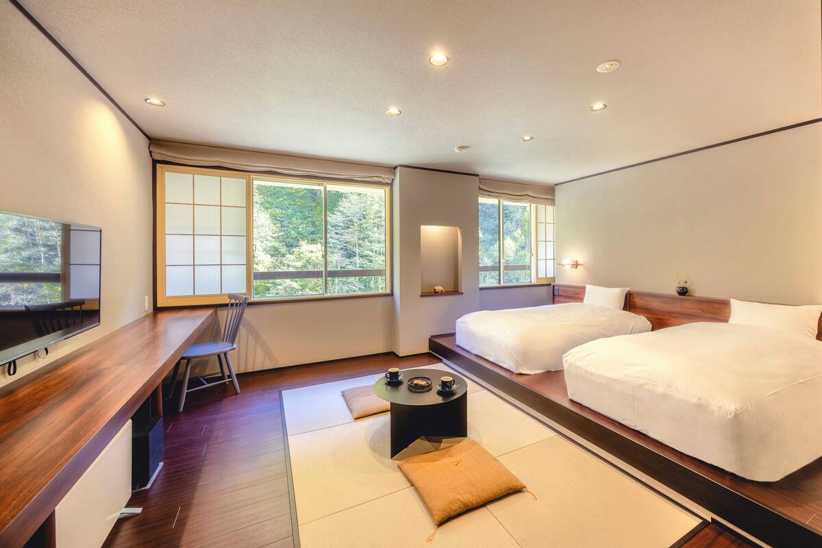 Une chambre d’hôtel moderne avec un bureau en bois, deux lits, une télévision et de grandes fenêtres donnant sur la verdure.