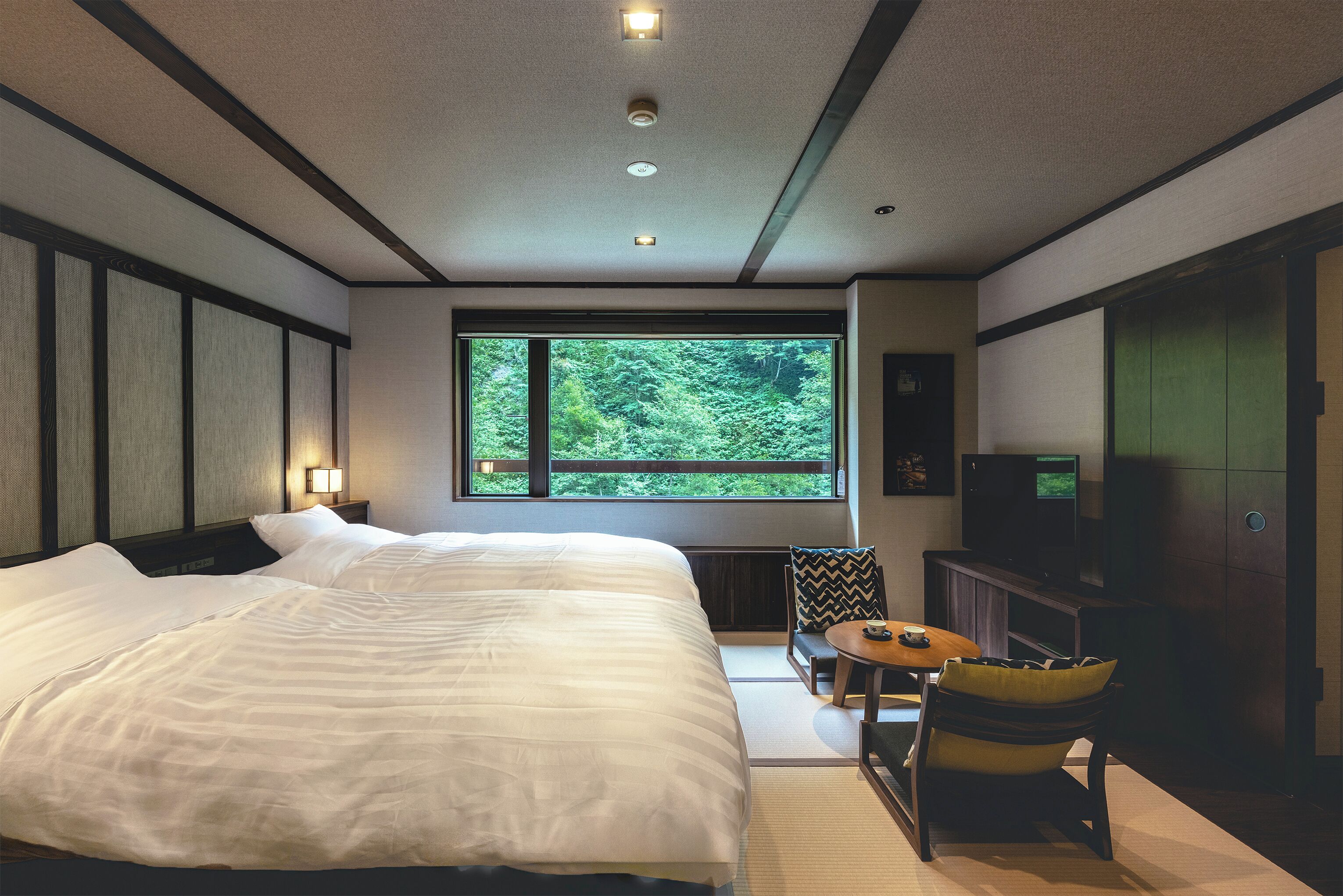 A hotel room with a large bed, a television, a small table, and a view of greenery through a window.