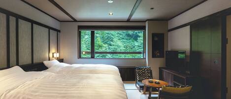 A hotel room with a large bed, a television, a small table, and a view of greenery through a window.