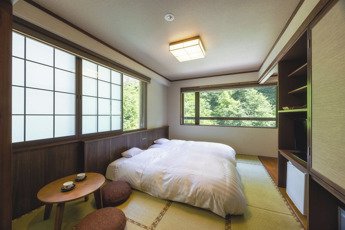 Une pièce de style japonais traditionnel, avec un sol recouvert de tatamis, un grand lit, une petite table avec deux tasses et un téléviseur posé sur une étagère.