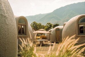 A series of dome-shaped accommodations with arched windows, a central table with an umbrella, and a natural landscape in the background.