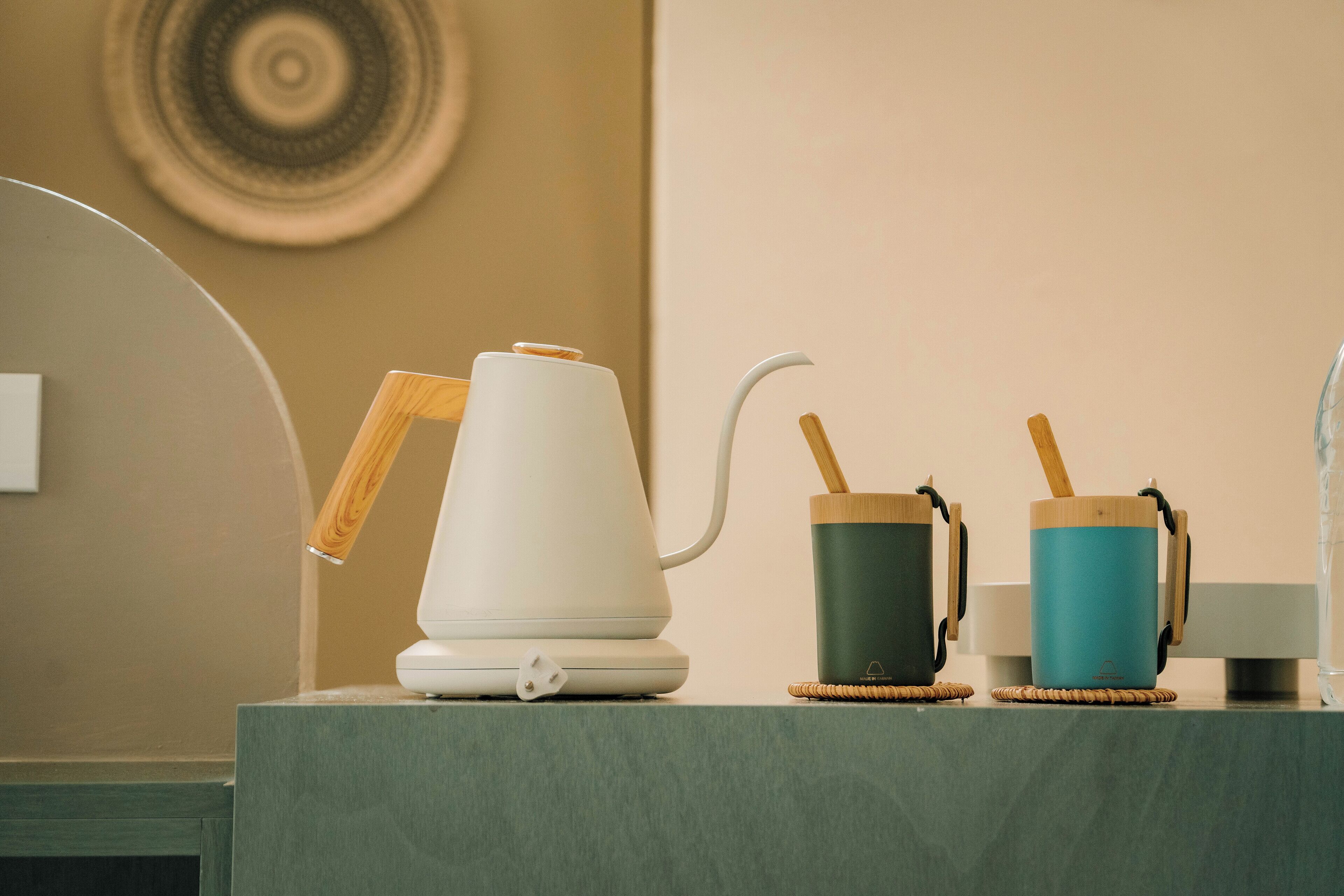 A kitchen counter with a white electric kettle, a wooden spoon, and two wooden cups on woven coasters.