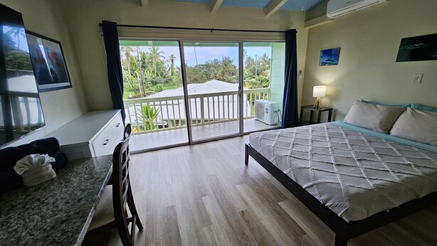 Une chambre avec un lit, un bureau, une chaise, un balcon donnant sur des palmiers et un appareil de climatisation.