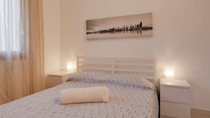 Una cama bien hecha con una colcha estampada y una manta doblada, un cuadro de un paisaje urbano en la pared, y lámparas de noche sobre una mesita blanca.