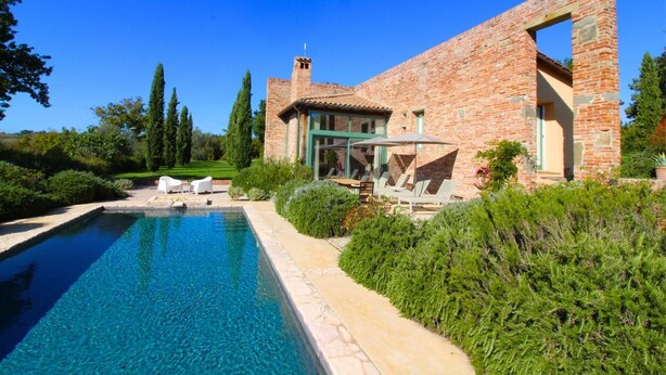 Una gran piscina con cubierta de piedra, una casa de ladrillo con invernadero de vidrio y un jardín bien cuidado con árboles y arbustos altos.