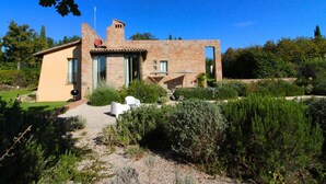 Una casa de piedra rústica con una construcción anexa pequeña, rodeada de abundante vegetación y un camino de grava.