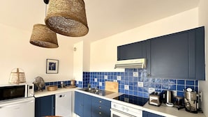 Une cuisine de taille modeste avec des armoires bleues, une cuisinière blanche et un four à micro-ondes. On y trouve des luminaires en osier au plafond, une table en bois et un tableau encadré au mur.