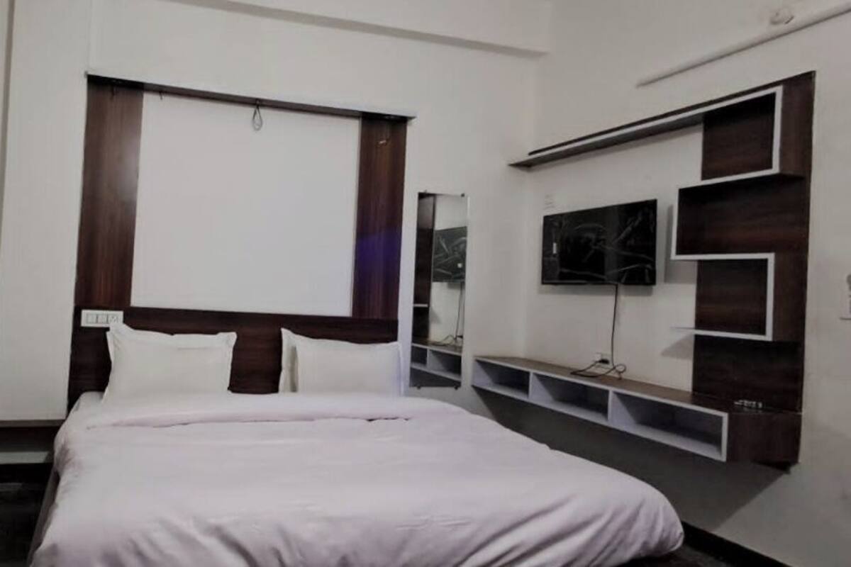 Una habitación de hotel con cama, mesita de noche, televisor y estante en la pared.