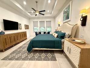 Une chambre à coucher comprenant un lit, un ventilateur de plafond, une télévision, une armoire en bois et une fenêtre avec des stores.