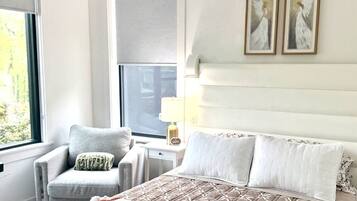 Un dormitorio con cama, una silla, un ventanal y dos cuadros en la pared.