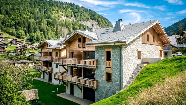 A modern chalet with a stone facade, wooden balconies, and a view of a mountainous village.
