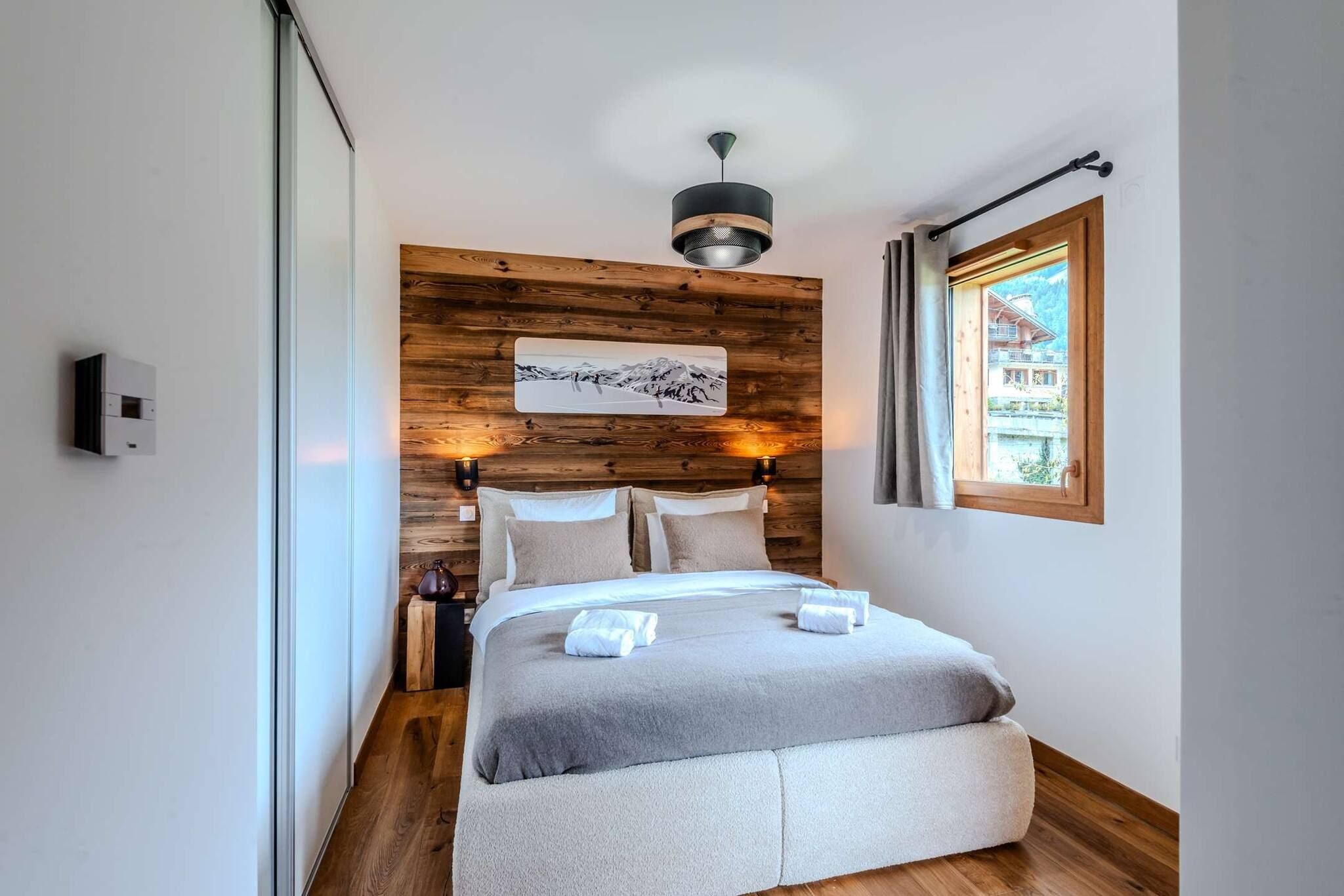 Ett modernt sovrum med en sänggavel i trä, en säng med grått sänglinne, ett upphängt konstverk, ett nattduksbord med en lampa och ett fönster med utsikt över en byggnad.