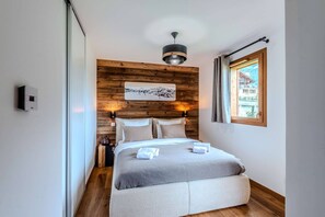 Een moderne slaapkamer met een houten hoofdbord, een bed met grijze beddengoed, een hangend kunstwerk, een nachtkastje met een lamp, en een raam met uitzicht op een gebouw.