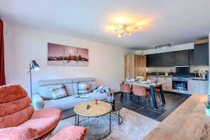 A modern living room with a sofa, a coffee table, and a dining area with chairs. There is a kitchen with dark cabinets and a stove. - Charming Apartment – Ideal for skiing and MTB (MORZINE)