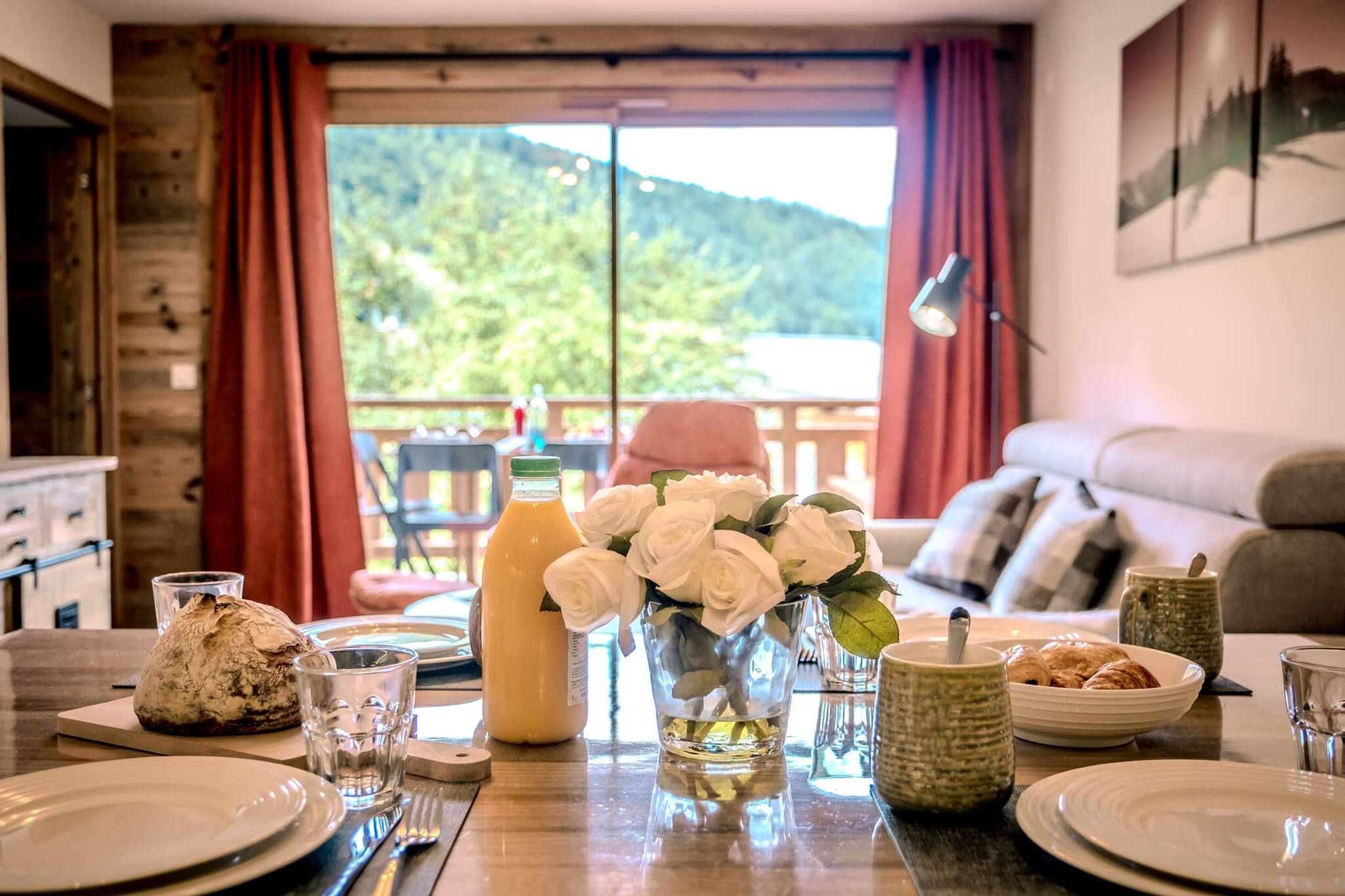 A dining table set with plates, glasses, a loaf of bread, a bottle of juice, a vase with white flowers, and a plate of croissants.