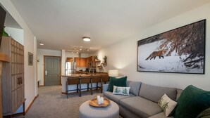 A living room with a grey sofa, a round ottoman, and a dining area with wooden cabinets and a kitchen in the background.