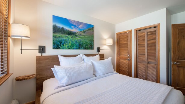 A bedroom with a large bed, two wall-mounted lamps, a wooden headboard, a painting of a landscape, and a set of wooden closets.