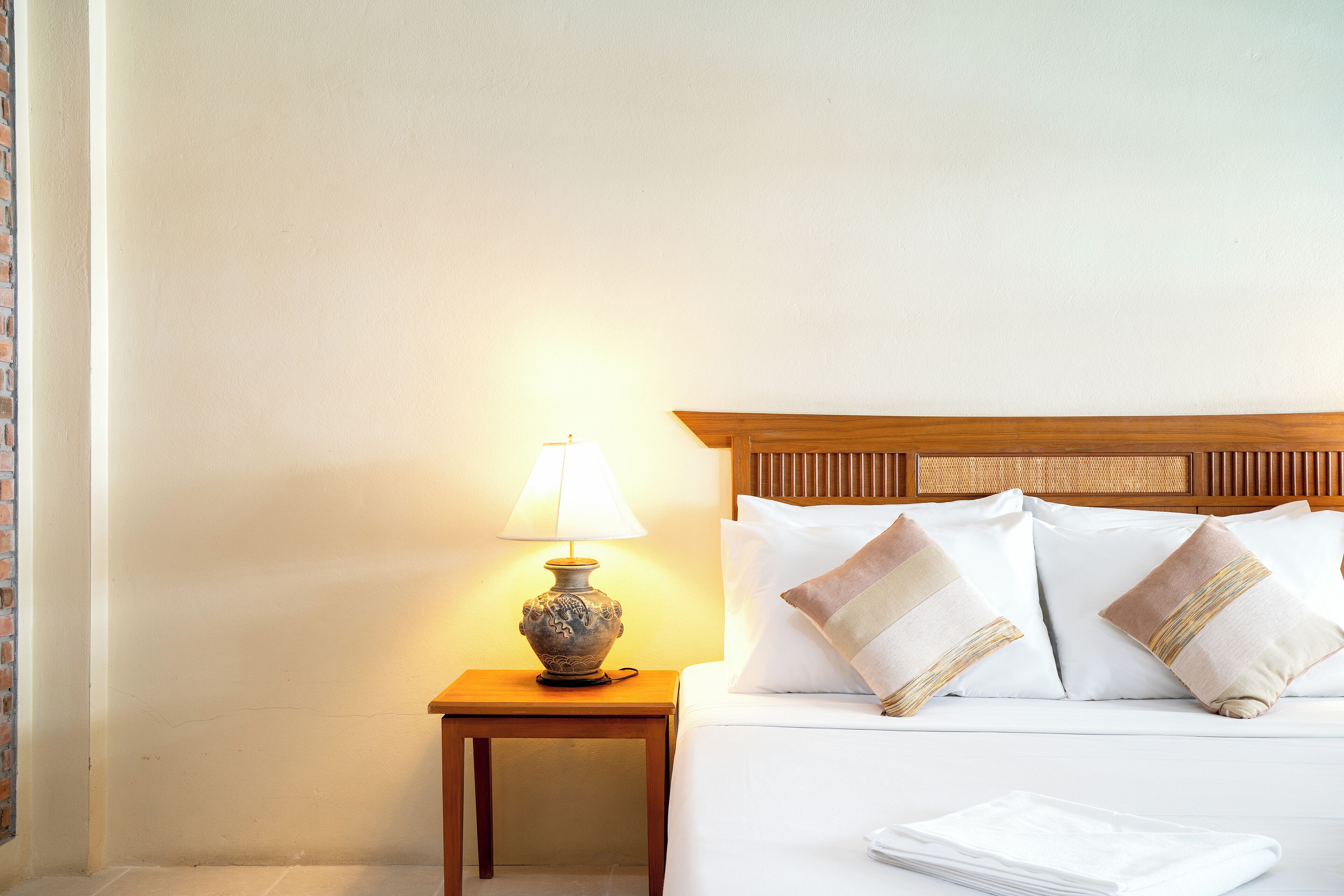 A neatly made bed with white linens and decorative pillows, a small wooden nightstand with a lamp, and a plain wall.