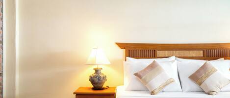 A neatly made bed with white linens and decorative pillows, a small wooden nightstand with a lamp, and a plain wall.