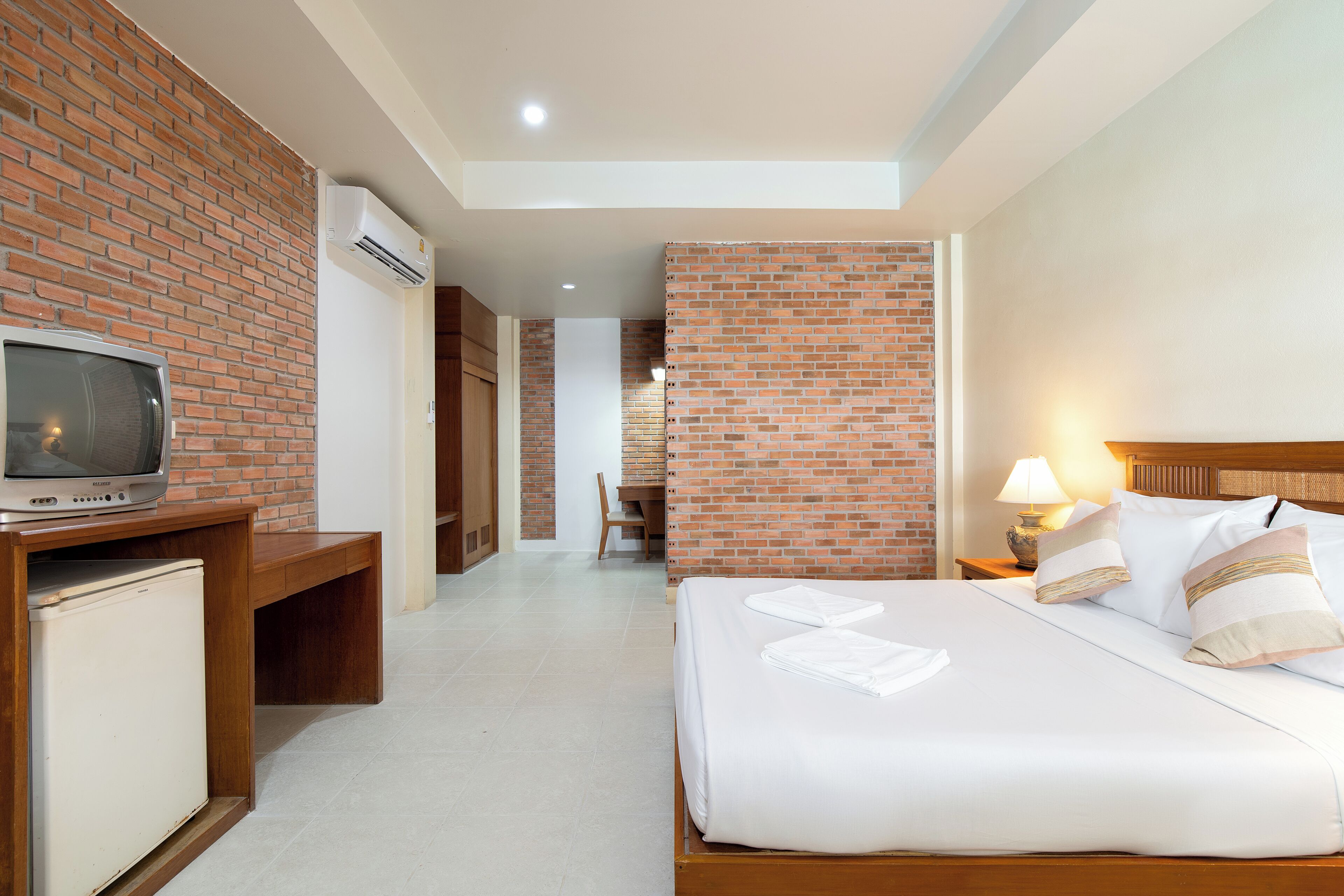 A hotel room with a brick wall, a television on a wooden table, a bed with white linens, and a nightstand with a lamp.