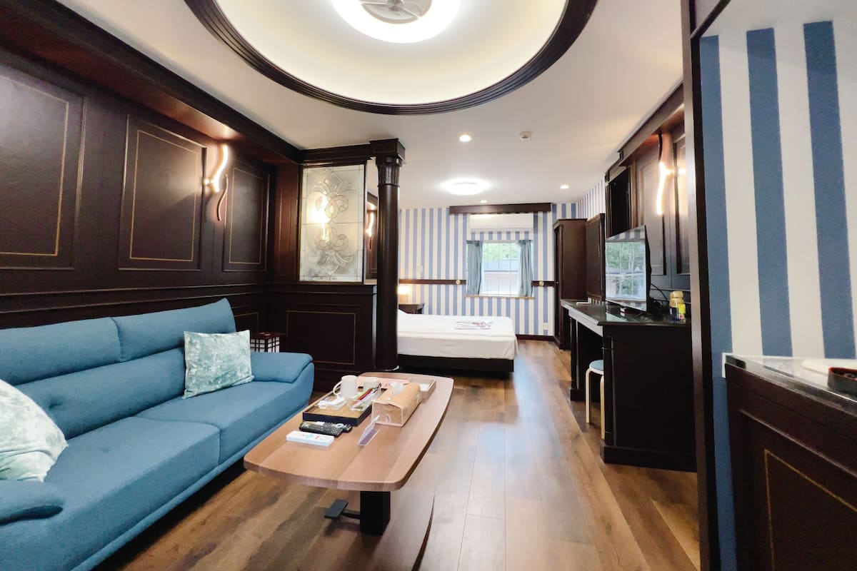 A hotel room with a blue sofa, a wooden coffee table, a bed, a TV, and a kitchen area.