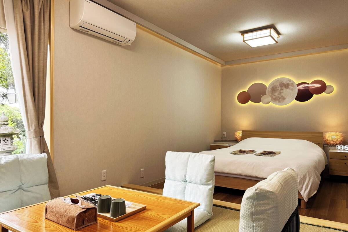 A hotel room with a bed, bedside tables, a wooden table with a bag and cups, and a wall-mounted air conditioner.