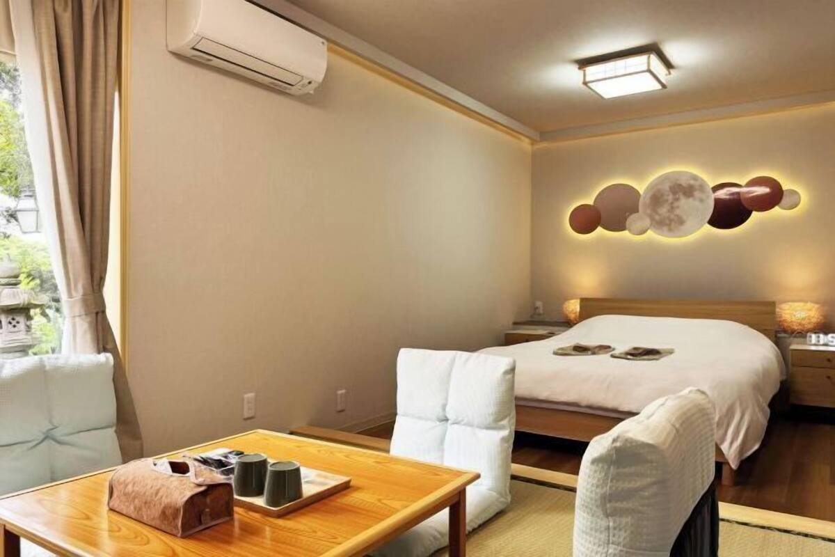 A hotel room with a bed, a bedside table with a lamp, a chair, a table with a bag and a tray, and a wall-mounted air conditioner.