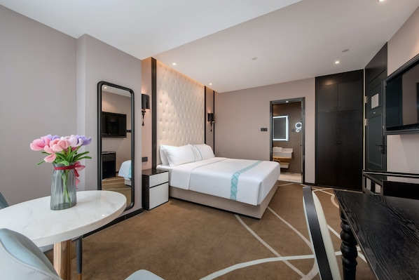 A hotel room with a bed, a desk, a chair, a mirror, a TV, and a vase with flowers.
