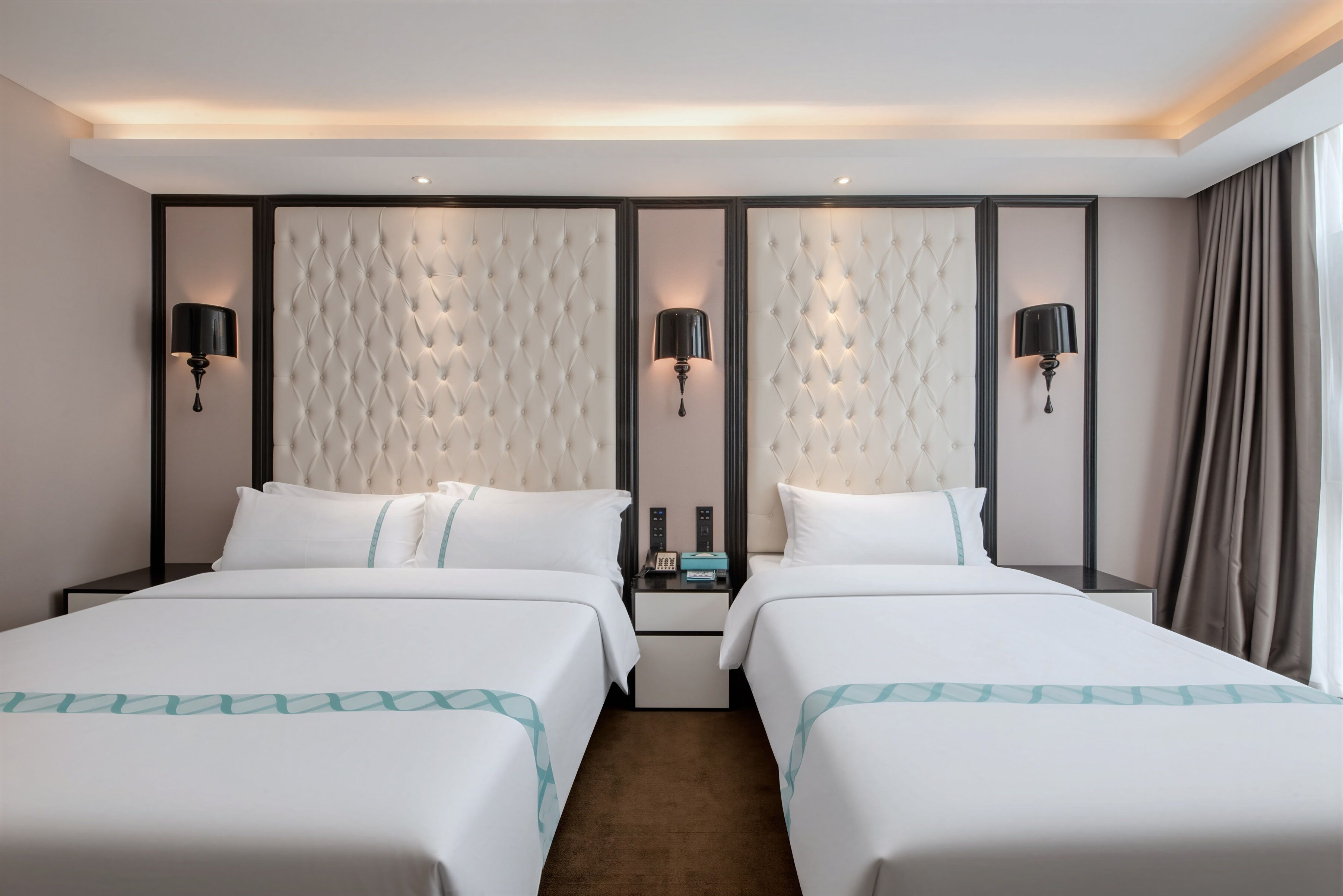 A hotel room with two beds, a tufted headboard, bedside lamps, a nightstand, and a window with curtains.