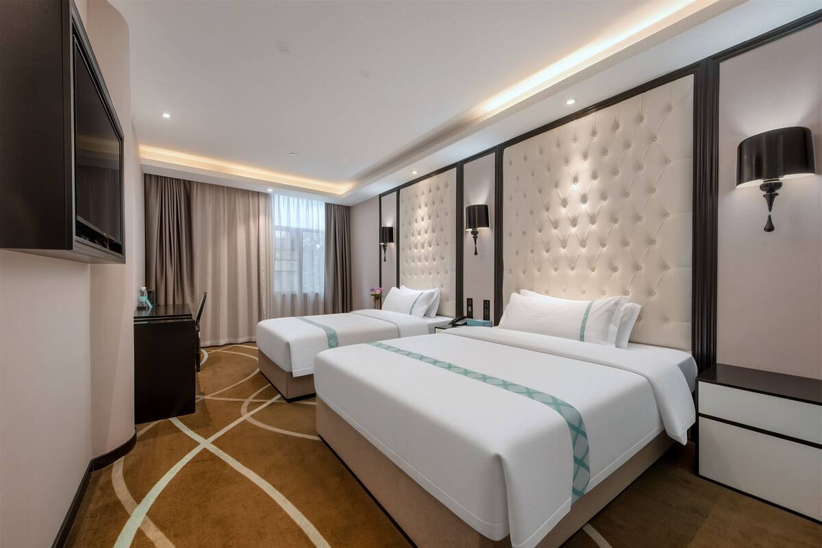 A hotel room with two beds, a flat-screen TV, a minibar, a large window with curtains, and a tufted headboard.