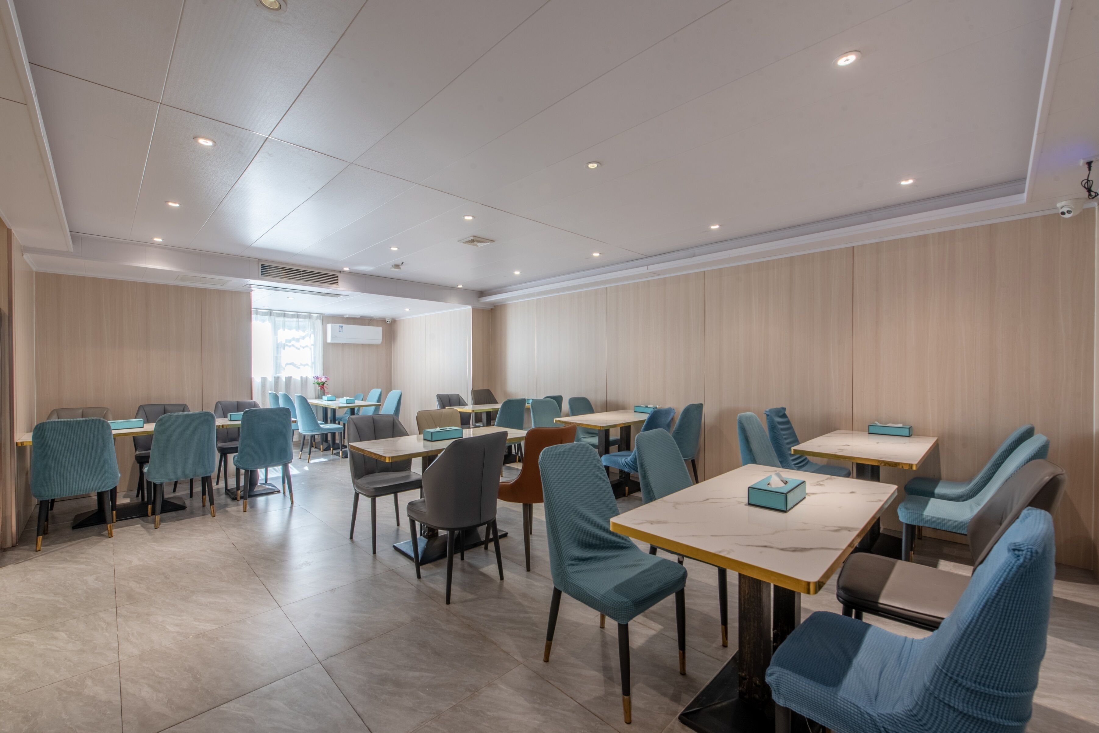 A spacious dining area with multiple tables and chairs, a ceiling with recessed lighting, and a view of the outdoors through a window.