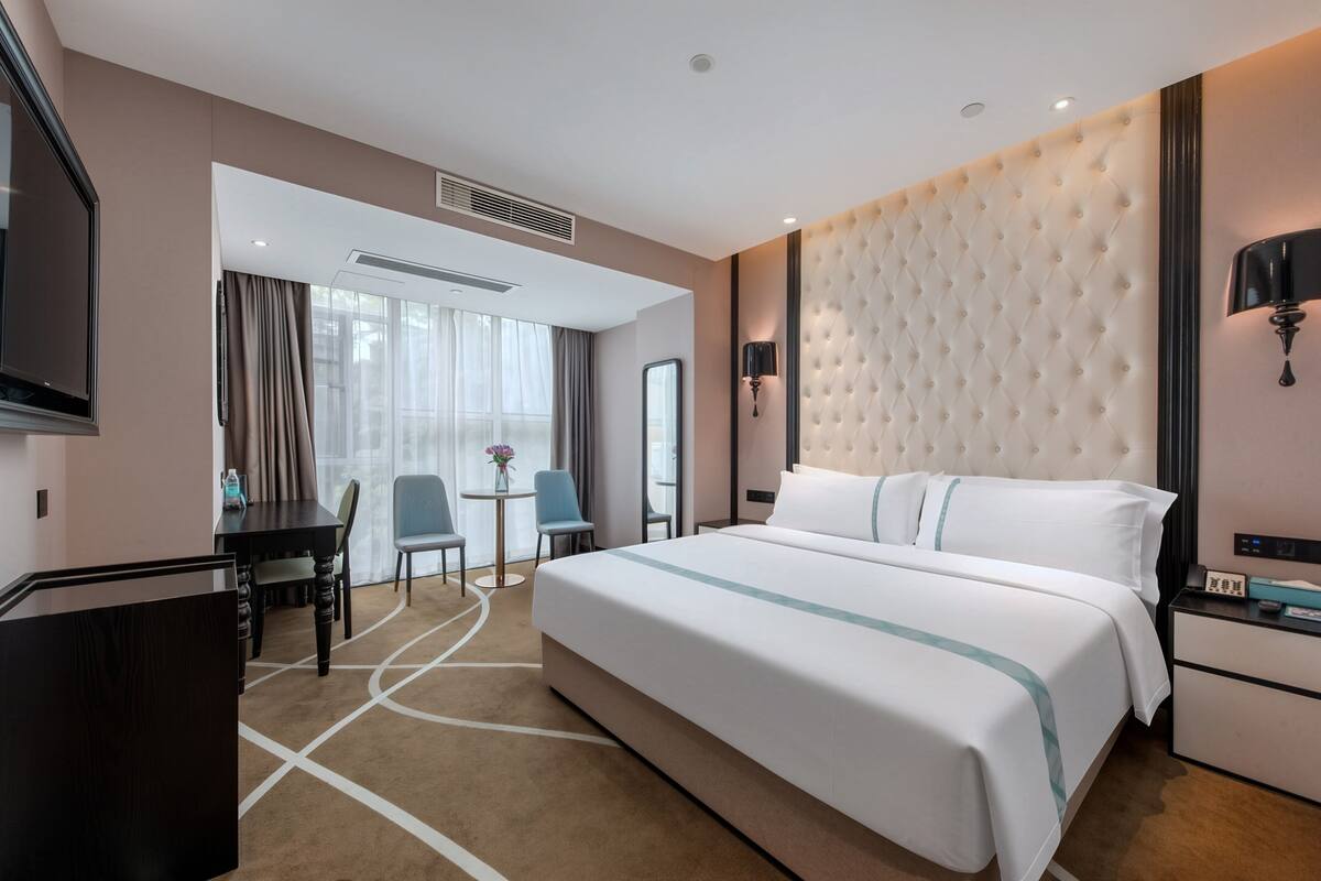 A hotel room with a large bed, a desk, a chair, a small table with a vase, and a TV.
