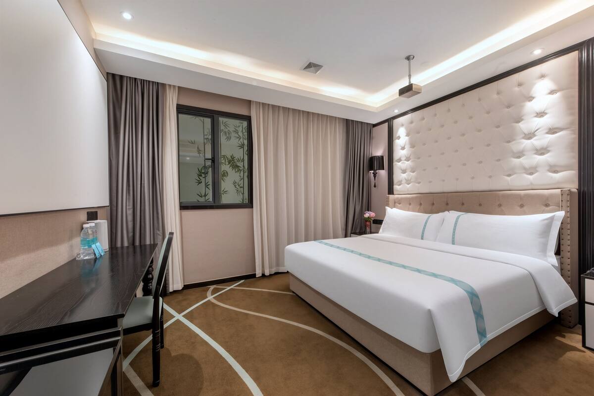 A hotel room with a bed, a desk, a chair, a window with curtains, a nightstand, and a lamp.