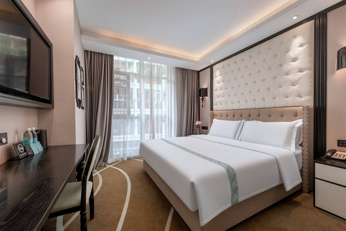 A hotel room with a large bed, a desk with a flat-screen TV, a bedside table, and a large window with curtains.