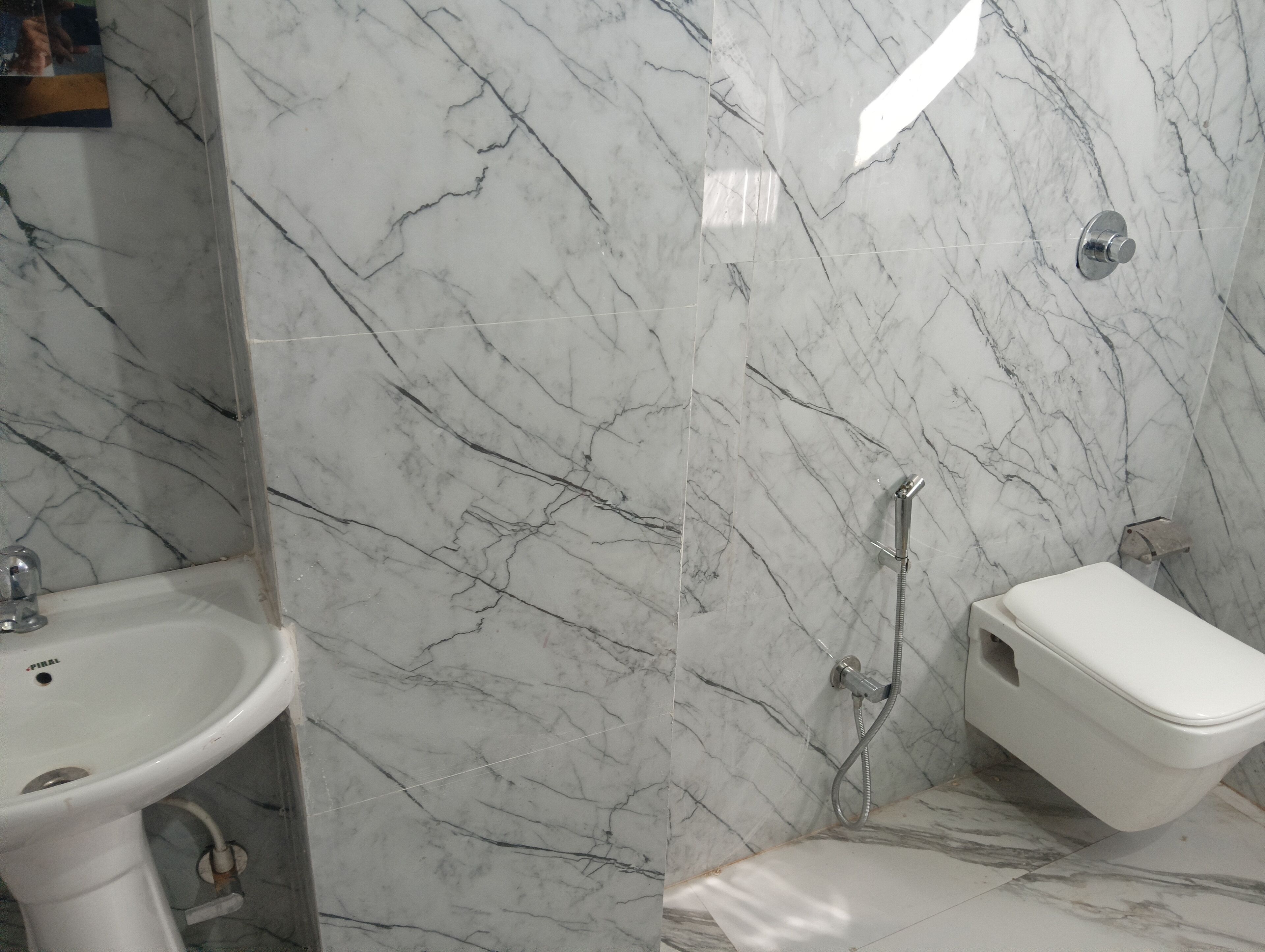 Baño con lavamanos, inodoro y zona de ducha blancos, todo con suelo y paredes de apariencia marmolada.
