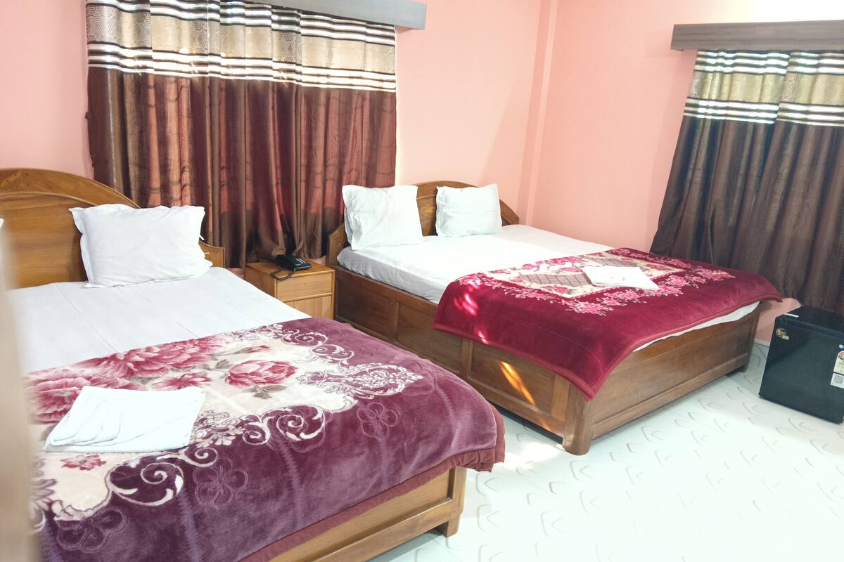 A hotel room with two beds, each with a wooden headboard, white bedding, and a floral-patterned blanket. There is a nightstand with a phone between the beds, and a small black suitcase is visible on the floor.