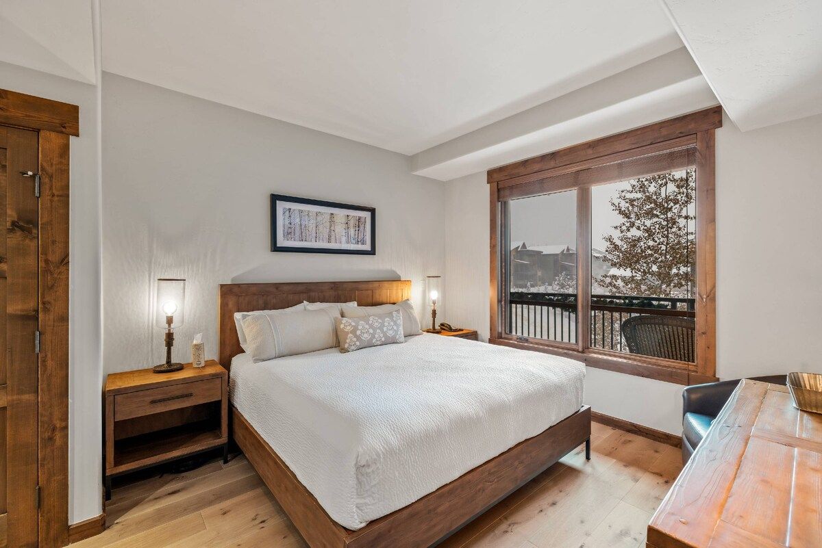 A bedroom with a large bed, wooden headboard, bedside tables with lamps, a window with a view, and a wooden floor.