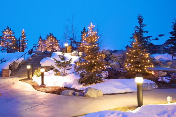 A snow-covered outdoor area with illuminated Christmas trees and a staircase leading up to a building.