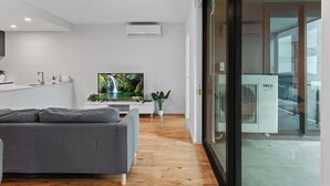 Een moderne woonkamer met een grijze bank, een flatscreen televisie, een klein wit tafeltje en een groot raam.