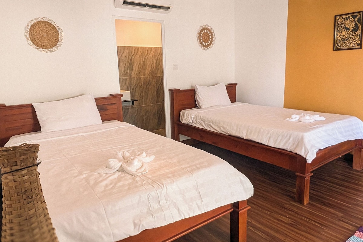 A hotel room with two wooden beds, a wooden bench, a woven basket, and a wall-mounted air conditioner.